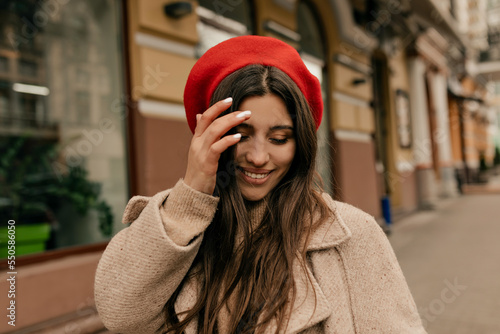 Close up portrait of lovely cute woman touching her hair, looking down and smiling. She wears red beret, beige coat  © PhotoBook