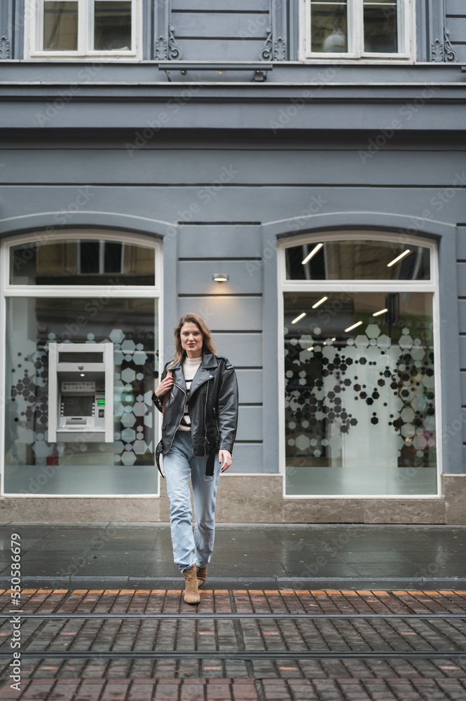 Young street fashion blond woman crossing the street in urban city environment, looking at camera, black leather jacket and blue jeans, elegant boots. Full length portrait, autumn collection. Young street fashion blond woman crossing the street in urban city environment, looking at camera, black leather jacket and blue jeans, elegant boots. Full length portrait, autumn collection.