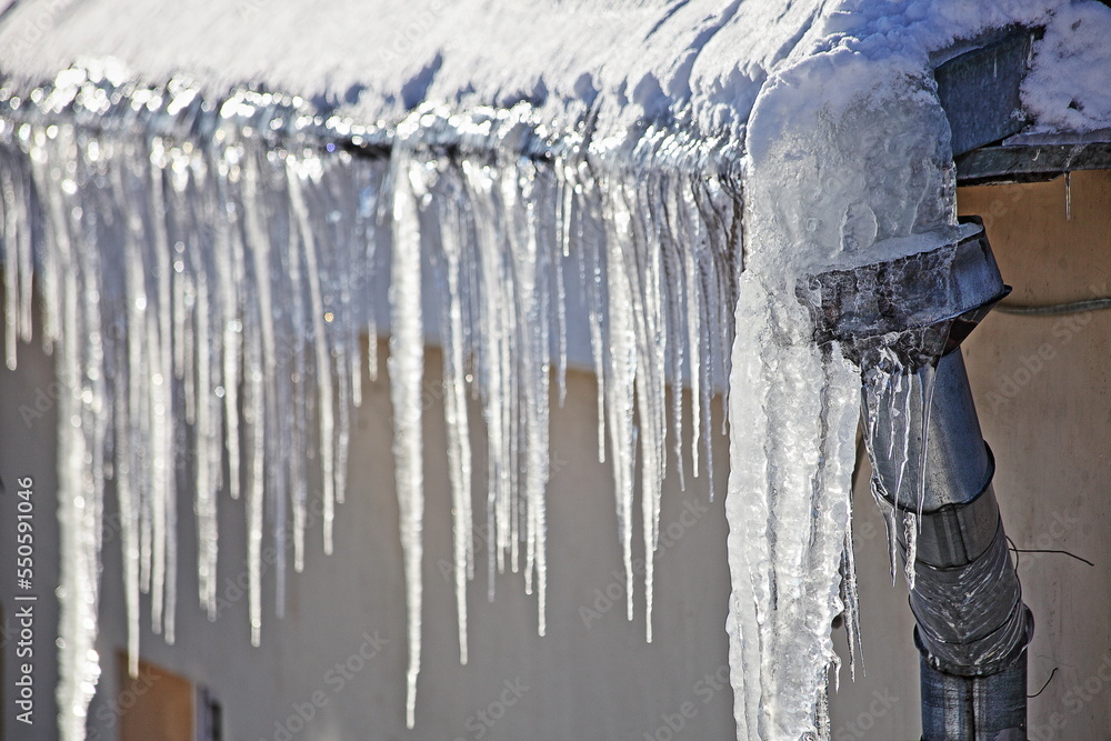 Big icicles on snow covered old roof with downpipe intake on city house at Sunny winter day in Europe