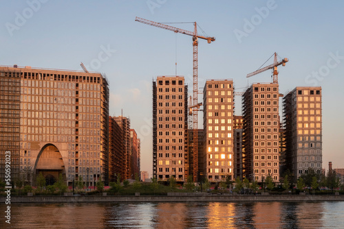 Construction site for residential complex at embankment with cranes and reflection in river.