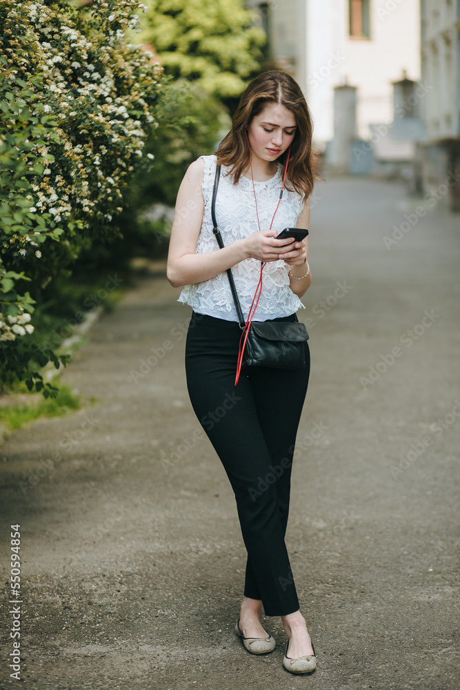 Fototapeta premium Happy beautiful young Caucasian girl with smart phone outdoors on sunny summer day texting and smiling
