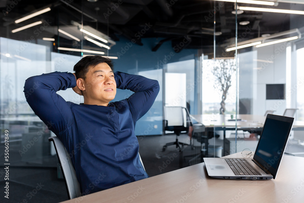 Asian man finished work well, businessman resting in office satisfied with achievement throw hands behind head dreaming, man working inside office using laptop at work. Asian man finished work well, businessman resting in office satisfied with achievement throw hands behind head dreaming, man working inside office using laptop at work.
