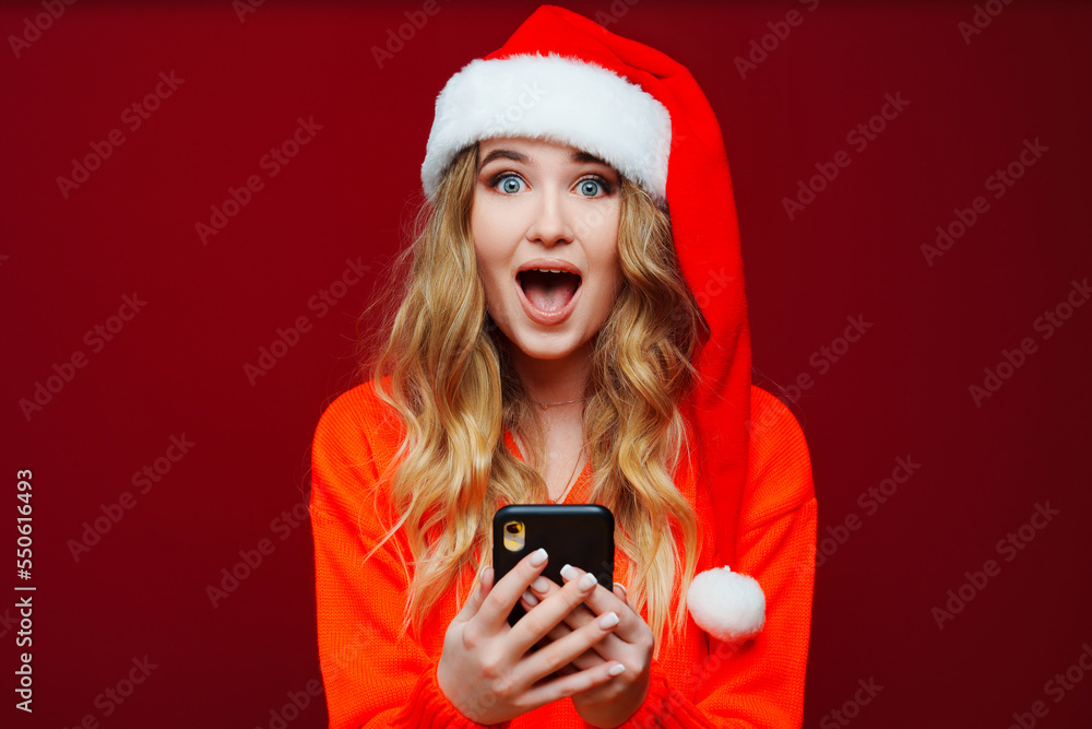 a surprised woman in a Santa Claus hat with smartphone on a red background. a surprised woman in a Santa Claus hat with smartphone on a red background.