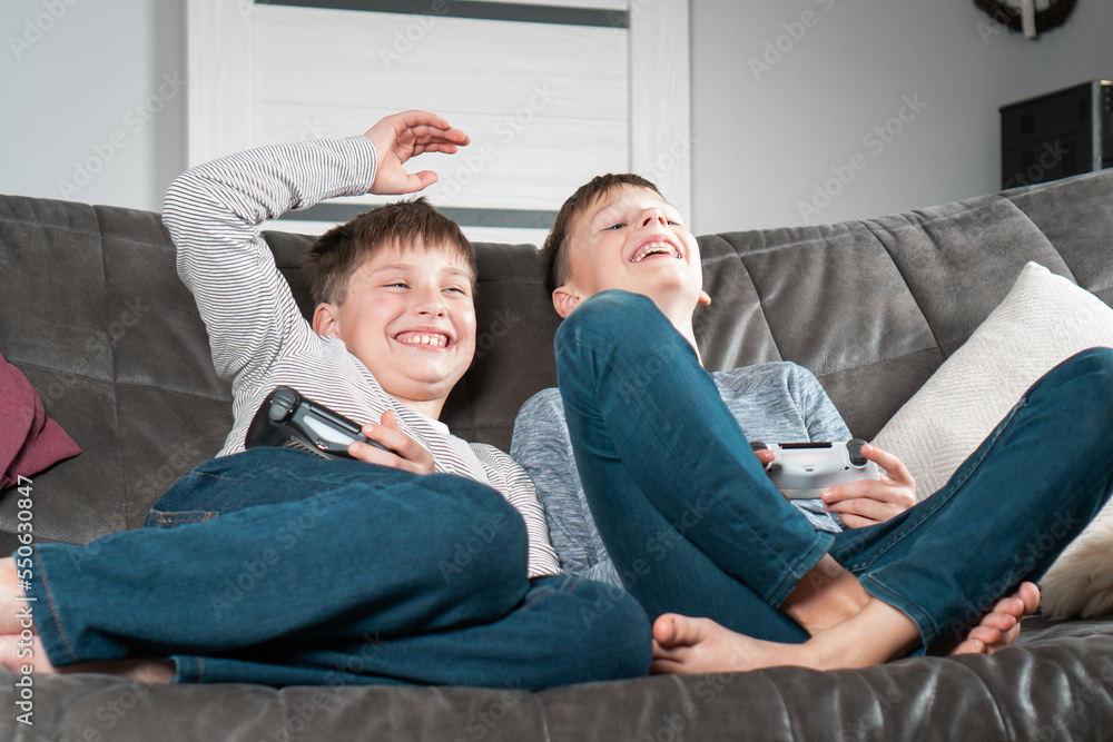 Laughing school aged boys sit on sofa with game joysticks. Children play videogames and have fun at home. Winning game.