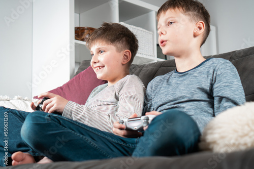 School aged boys sit on sofa with game joysticks, from below view. Children play videogames. Best friends lifestyle.
