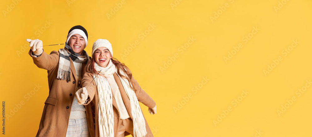 Portrait of stylish young man and woman cheerfully posing in winter clothes with sparkles isolated over yellow background. Flyer