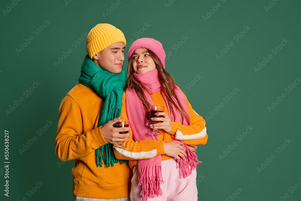 Portrait of young man and woman, lovely couple in bright knitted hat and scarf posing with mulled wine isolated over green background