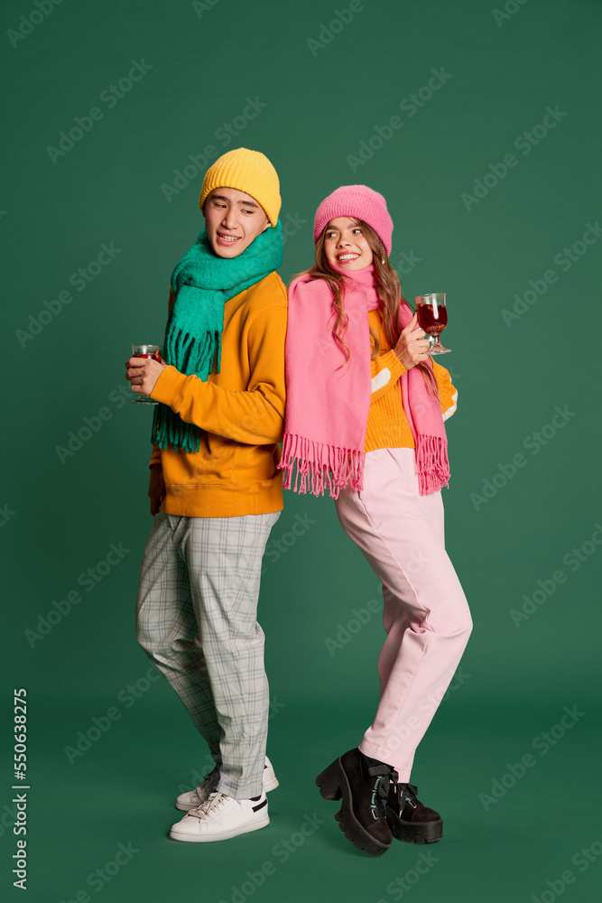 Portrait of young man and woman in bright knitted hat and scarf posing, drinking mulled wine isolated over green background Portrait of young man and woman in bright knitted hat and scarf posing, drinking mulled wine isolated over green background
