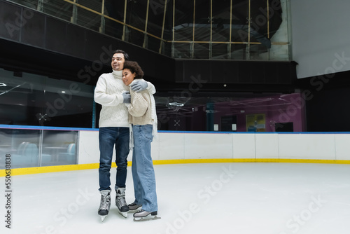 Smiling man hugging african american girlfriend in sweater and scarf on ice rink