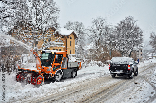 Straße im Winter mit Schneefall und Räumfahrzeug auf Gehweg