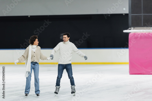 Smiling interracial couple holding hands and looking at each other while ice skating on rink