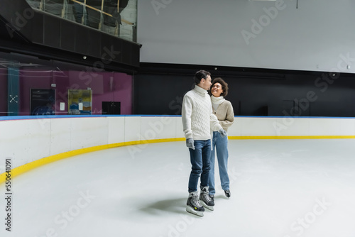 Positive multiethnic couple in warm sweaters ice skating on rink