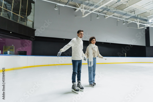 Happy interracial couple holding hands while ice skating on rink