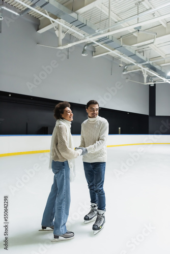 Positive multiethnic couple holding hands and looking away on ice rink