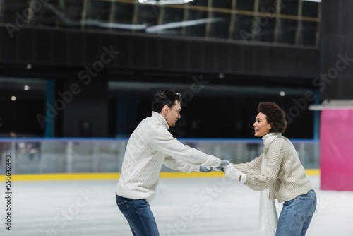 Side view of young interracial couple holding hands while ice skating on rink