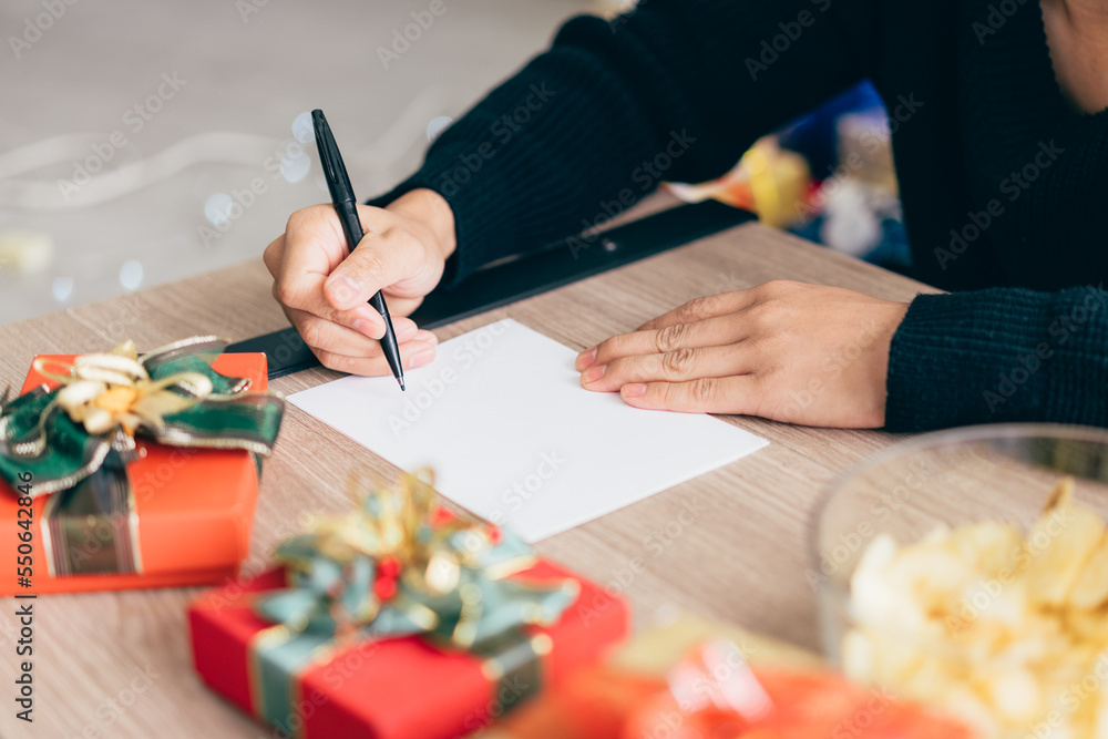 Close up man's hand is writing on a blank Christmas postcard with a pen. Couple sitting and writing Christmas card together for sending with surprise gifts at home during Christmas holiday.