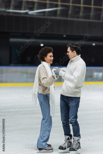 Young man in sweater holding hands of african american girlfriend in gloves on ice rink