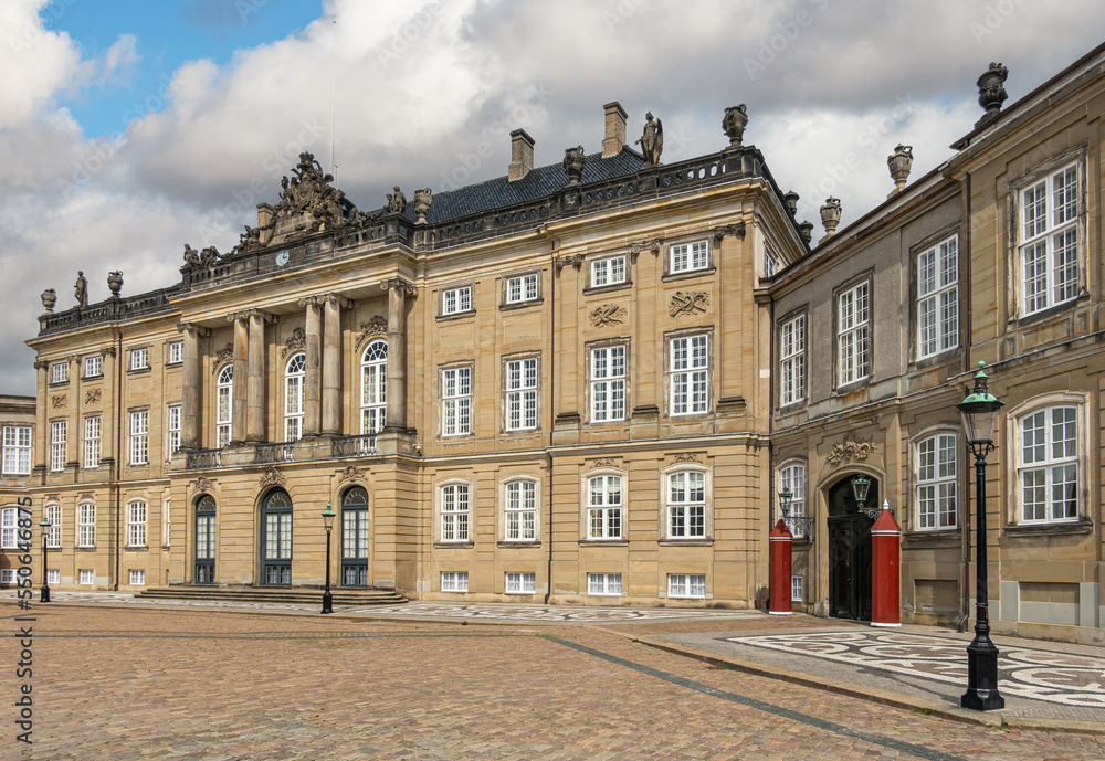 Copenhagen, Denmark - July 24, 2022: Brown stone Frederik VIII Palace and extra wing on Amalienborg under blue cloudscape. Brown stones with pediment statues, columns and classical architecture