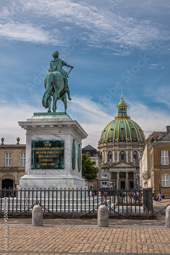 Copenhagen, Denmark - July 24, 2022: Greenish bronze Equestrian statue on pedestal of King Frederik V looking at his church in center of Amalienborg square under blue sky.