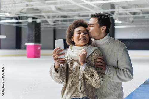 Young man kissing african american girlfriend taking selfie on ice rink