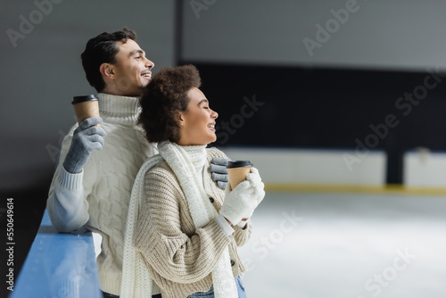 Side view of cheerful interracial couple holding coffee to go and looking away on ice rink
