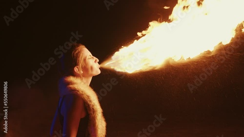 Young woman performing fire breathing using burning torch at night. Fire show artist lets out fiery cloud from mouth in open-air. Fire breather girl ignites flame at evening street show. Profile view photo