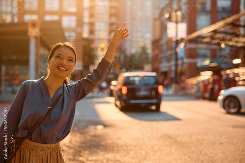 Murais de parede Happy Asian woman with hand raised stopping taxi in city.