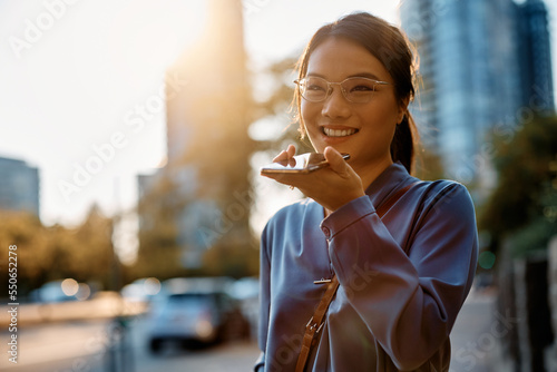 Happy Asian businesswoman talking over cell phone's speaker while walking in city.