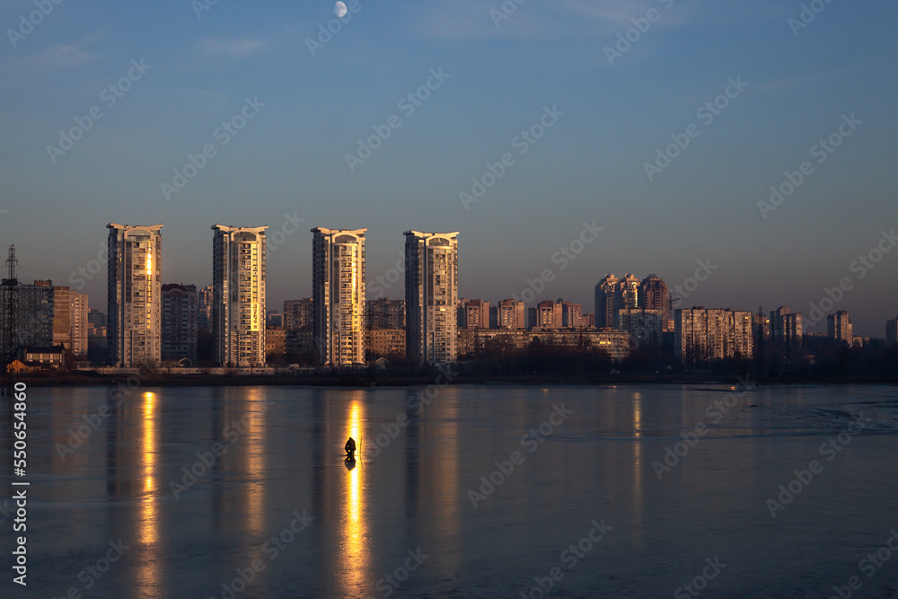 Obraz premium Fisherman on the frozen lake with skyscrapers in the background at the dusk