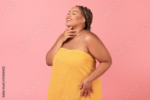 Excited african american body positive lady wrapped in towel touching her soft skin on neck, enjoying her young beauty