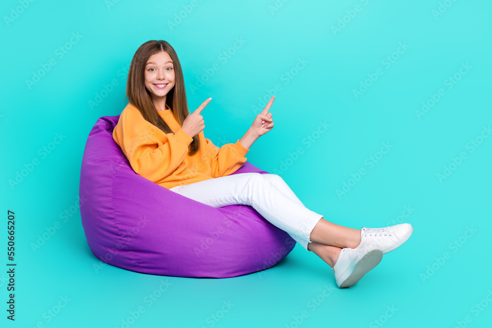 Full length photo of shiny funny schoolgirl wear orange sweatshirt bean bag two fingers empty space isolated turquoise color background Full length photo of shiny funny schoolgirl wear orange sweatshirt bean bag two fingers empty space isolated turquoise color background