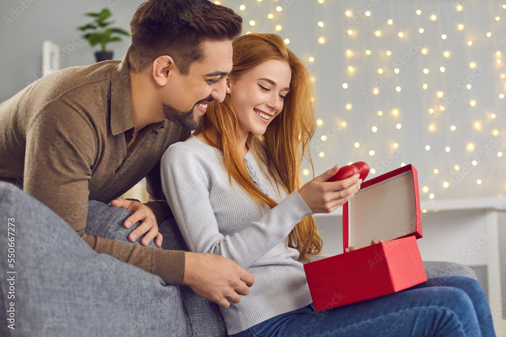 Smiling pretty young woman sitting and getting out red hearts from gift box present from her boyfriend for holiday at home. Love, togetherness, Christmas, birthday, valentines day, anniversary Smiling pretty young woman sitting and getting out red hearts from gift box present from her boyfriend for holiday at home. Love, togetherness, Christmas, birthday, valentines day, anniversary