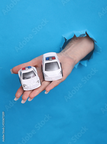 Female hand holds toy ambulance and police car through torn hole of blue paper