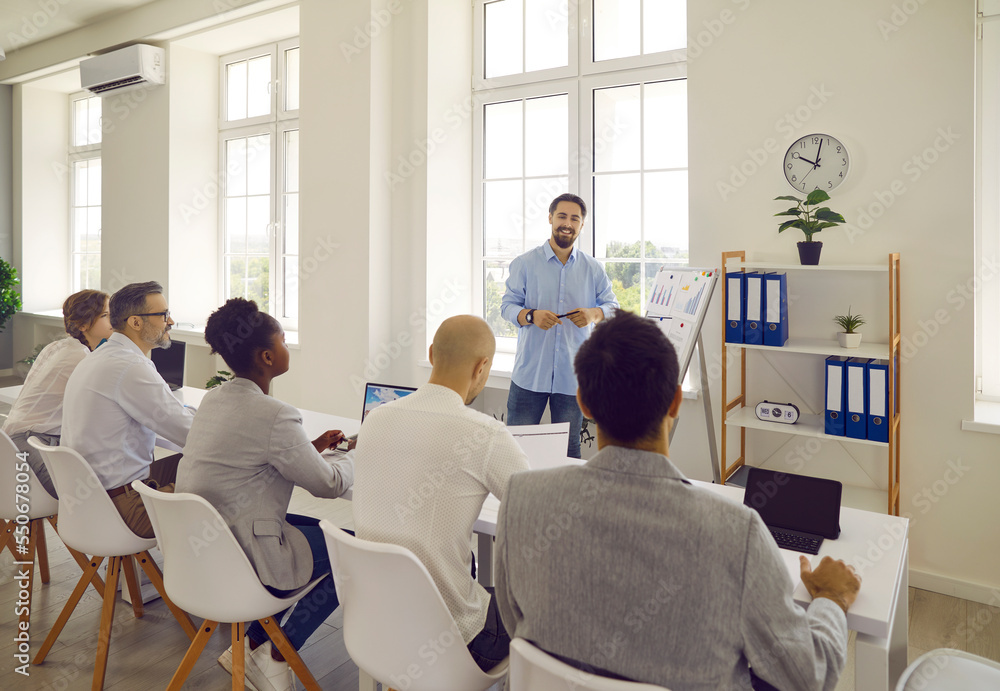 Obraz premium Man teacher business couch teaches communicates with a group of adult students in modern office. Students collegues sitting at desk back to camera. Corporative training, seminar, education, workshop.