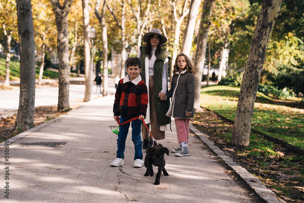Full body mom and kids with Labrador Retriever puppy on leash standing on asphalt path in sunlit autumn park in weekend