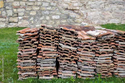 Old clay tiles are stacked on green grass, against background of old stone wall. Copy space. Selective focus.