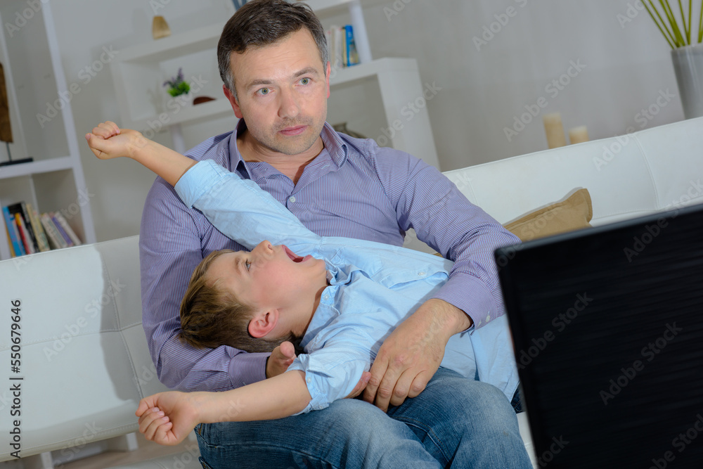 Fototapeta premium Man watching television, child layed across his lap yawning