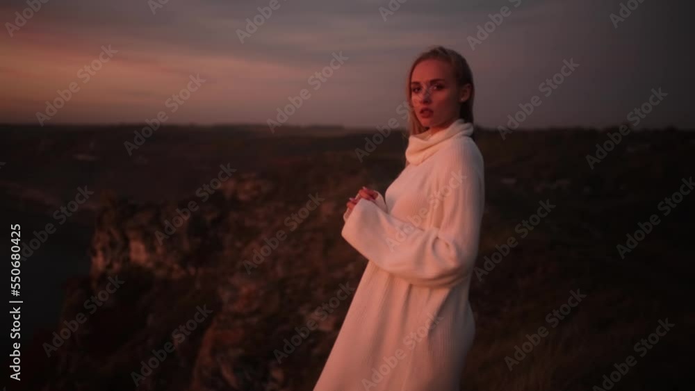 Portrait of a young beautiful blonde woman in a white knitted sweater on top of a mountain during an incredibly colorful sunset.