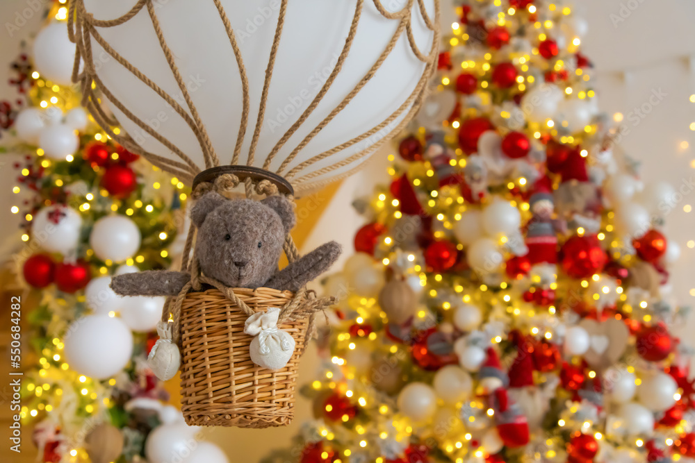 Small brown bear stuffed toy flies in basket on air balloon. Blurred Christmas tree in the background. Selective focus. Copy space for your text. Christmas travek theme.