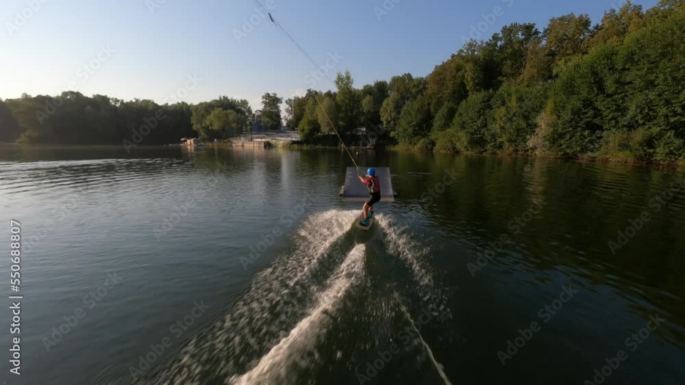 FPV drone flying following to female wakeboard surfer in helmet performing bad jump trick on ramp and falling into water at cable wake park, on background of sunlight at summer sunny day, slow motion. FPV drone flying following to female wakeboard surfer in helmet performing bad jump trick on ramp and falling into water at cable wake park, on background of sunlight at summer sunny day, slow motion.