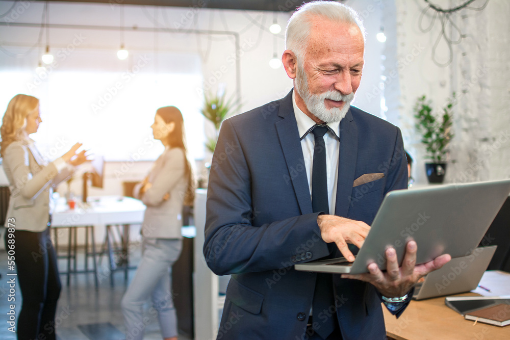 Portrait of senior businessman using laptop while standing in office. Portrait of senior businessman using laptop while standing in office.