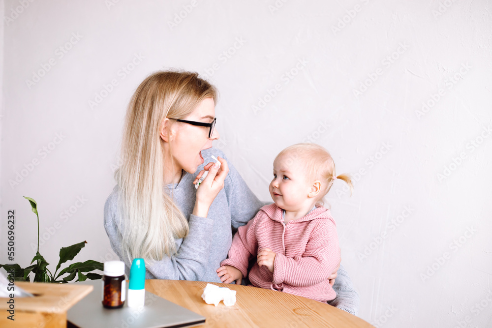 Side view of young mother woman sitting at table, holding little happy baby child, showing girl how to spray medicine.