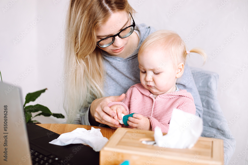 Portrait of young mother sitting at table near laptop, holding little baby, looking at bottle with medicine at home. Portrait of young mother sitting at table near laptop, holding little baby, looking at bottle with medicine at home.