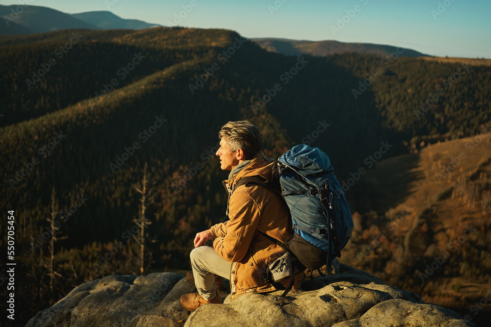 Confident alone man hiker with backpack sitting on stone of cliff and enjoying view of mountain summit. Active vacation hiking expedition outdoors