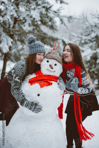 Close up fashion portrait of two sisters hugs and having fun, make snowman in winter time forest, wearing sweaters and scarfs,best friends couple outdoors, snowy weather