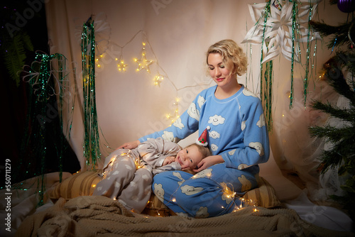 Cute mother and daughter in pajamas having fun in the room with Christmas garlands and white background. The tradition of decorating the house for the holidays. Happy childhood and motherhood