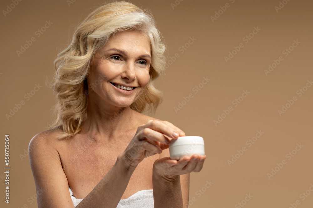 Attractive smiling middle aged woman holding jar of body cream isolated on beige background. Skin care, beauty products concept Attractive smiling middle aged woman holding jar of body cream isolated on beige background. Skin care, beauty products concept