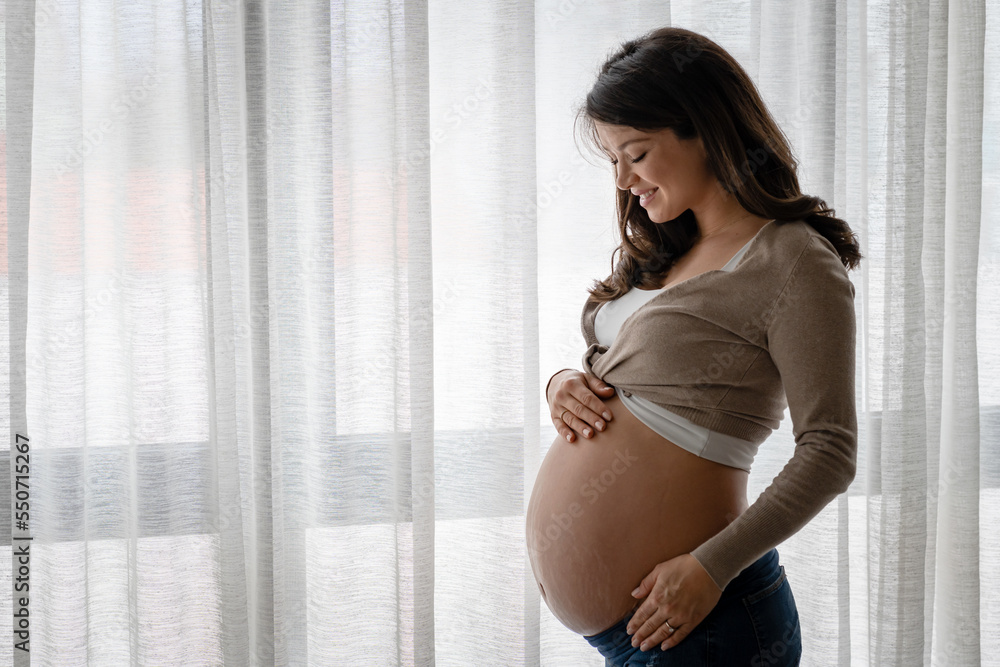 Portrait of a beautiful young pregnant woman standing by a big window and holding her belly. Always love your body. - obrazy, fototapety, plakaty 