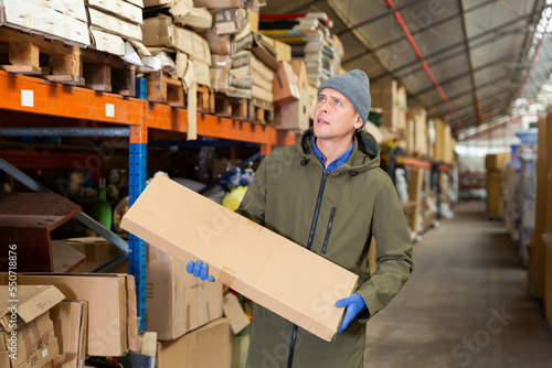 European man warehouse worker carrying pasteboard box during workday.