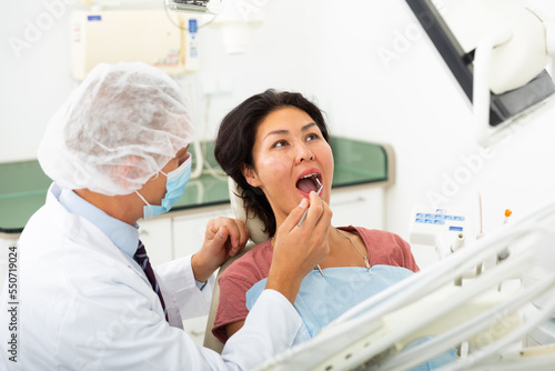 Qualified man dentist in a protective mask conducts an examines of a woman patient sitting in a dental chair in the clinic